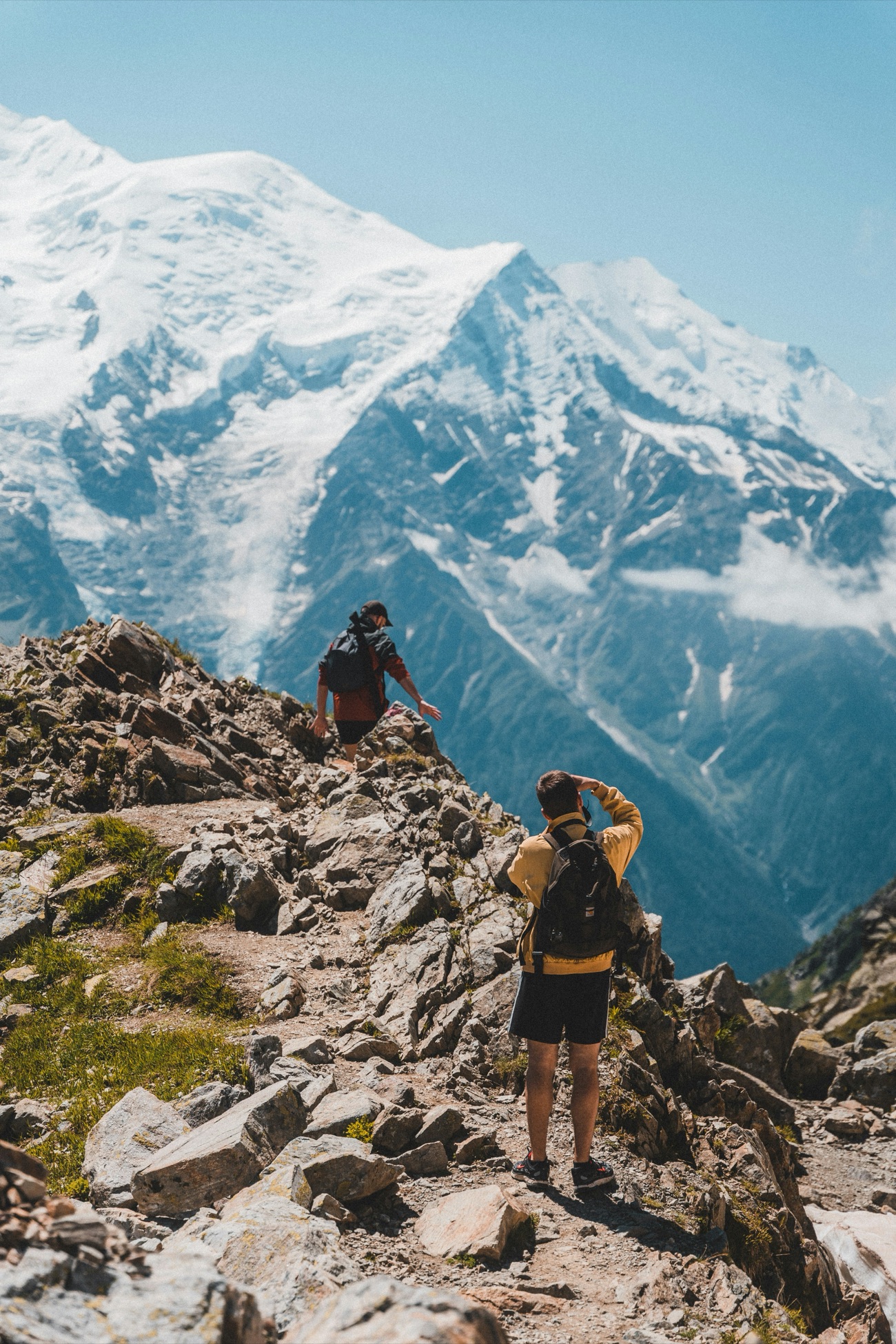 Two hikers climbing a rocky ridge toward a snow-capped peak.
