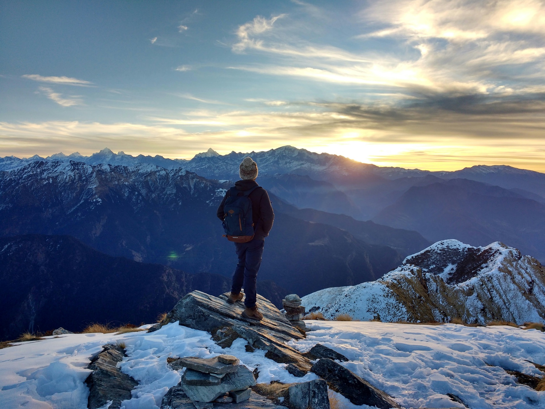 A hiker on a snowy ridge looking out over a mountain range at golden sunset.